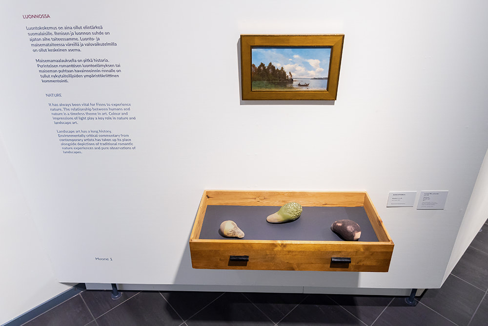 Three abstract and organic-shaped wooden sculptures exhibited in a wooden drawer attached on the wall, and above hangs a landscape painting